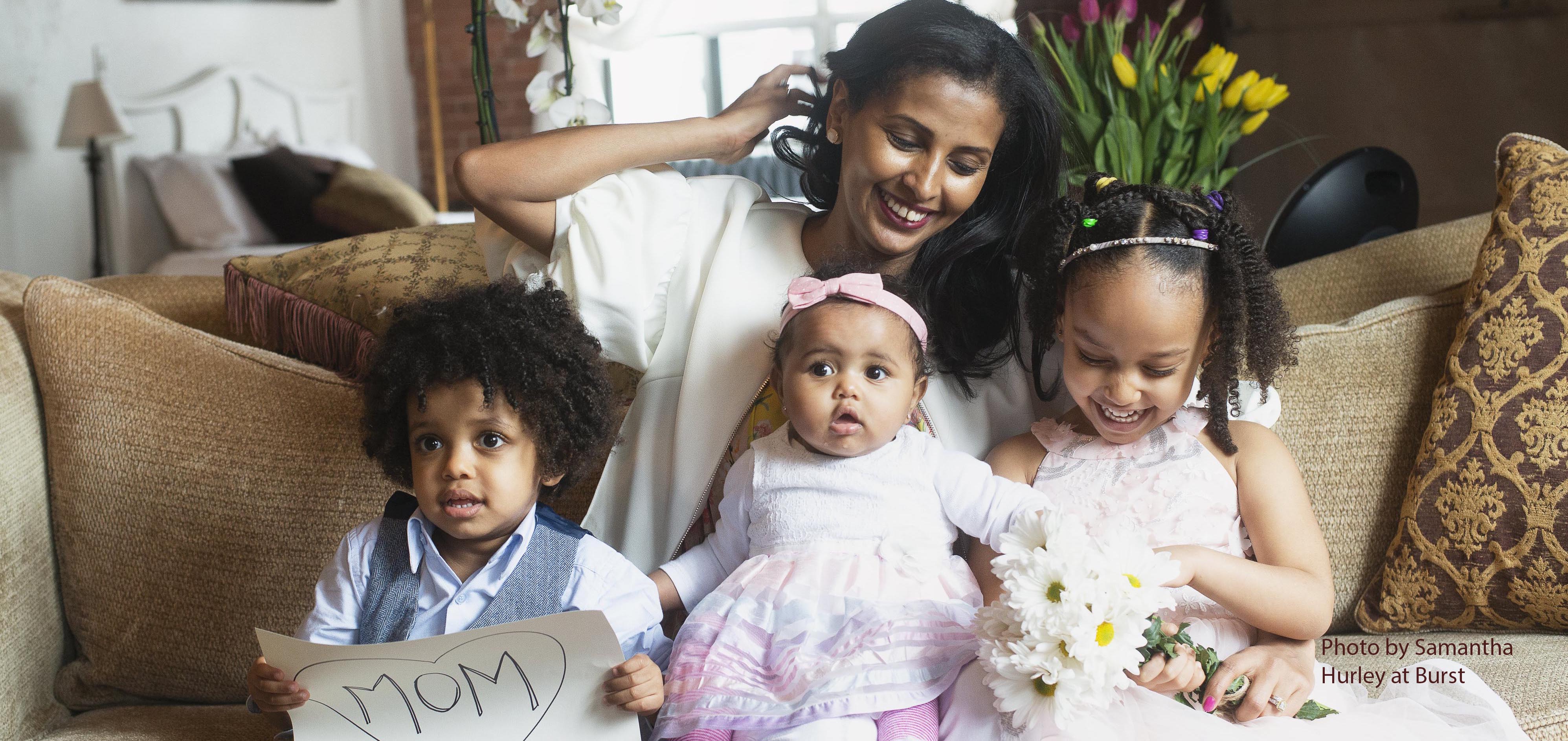 Woman on couch with three young children, one of whom holds a sign with "Mom" inside a heart