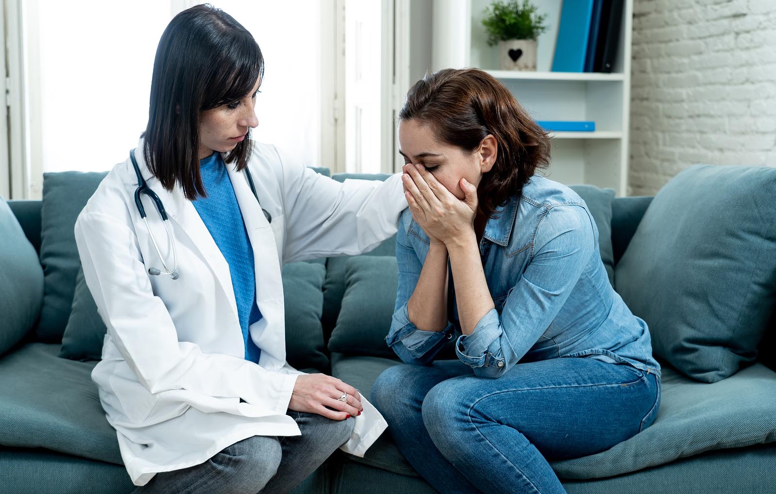 Woman in white coat with stethoscope rests her hand on the shoulder of woman with her head in her hands