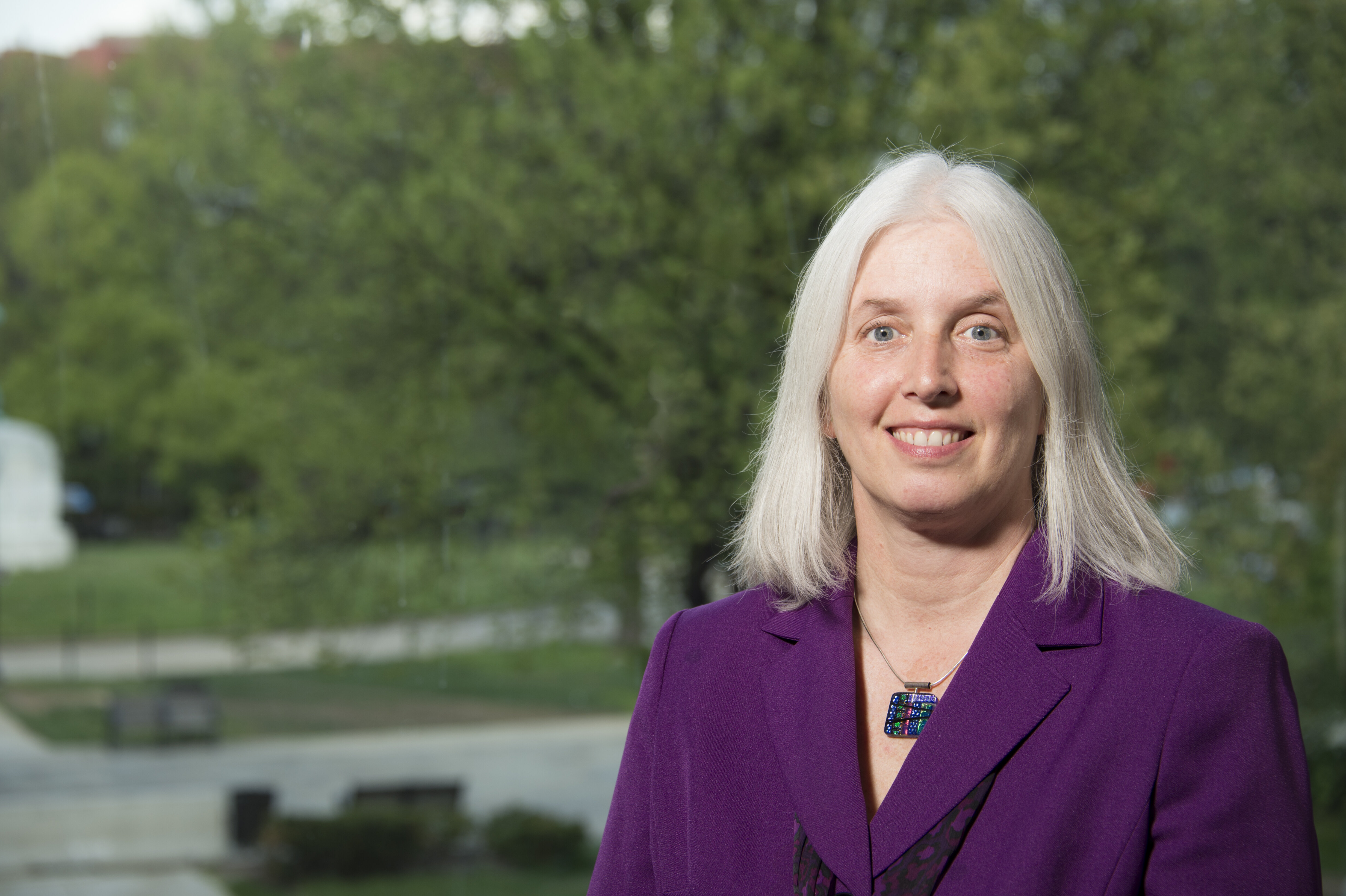 Susan Wood, a white woman with grey hair dressed in a purple suit, stands smiling in front of a tree