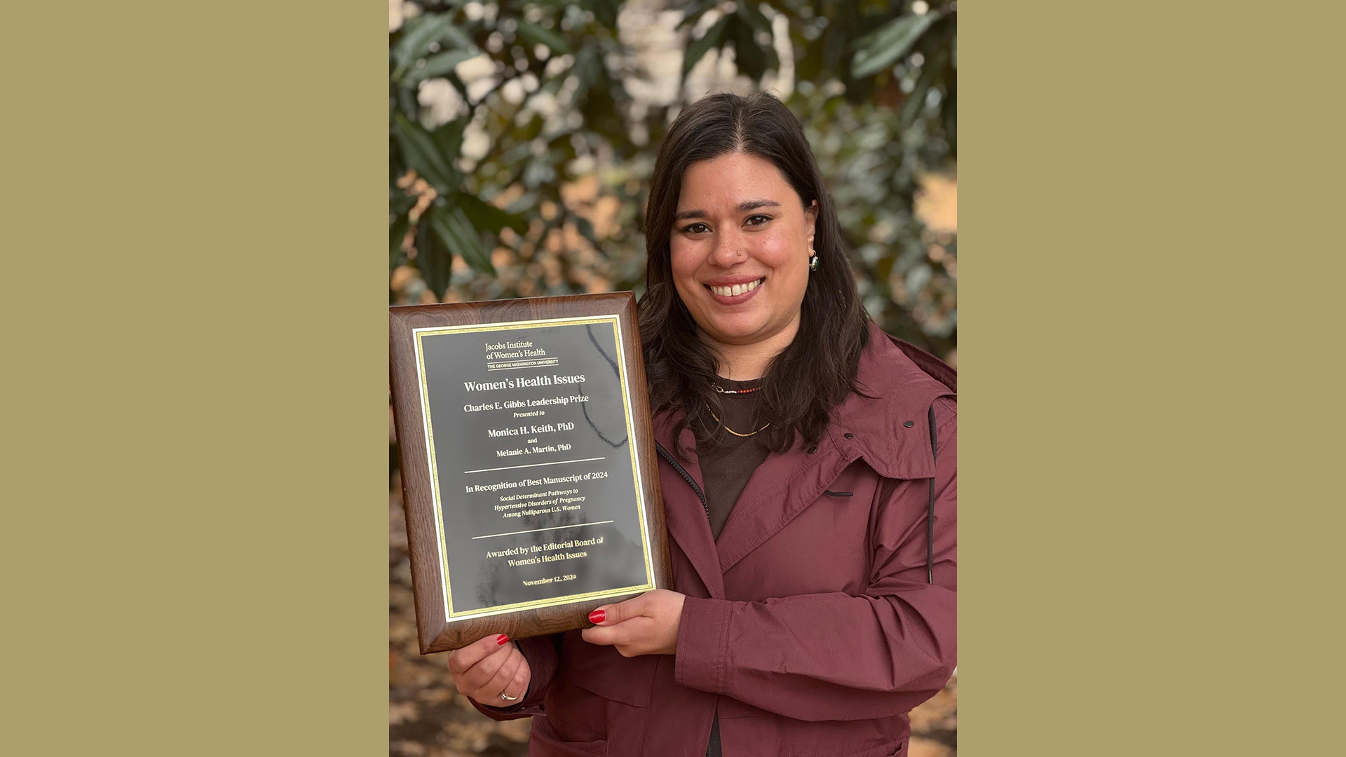 A smiling white-presenting woman with brown hair stands in front of a leafy background holding a plaque