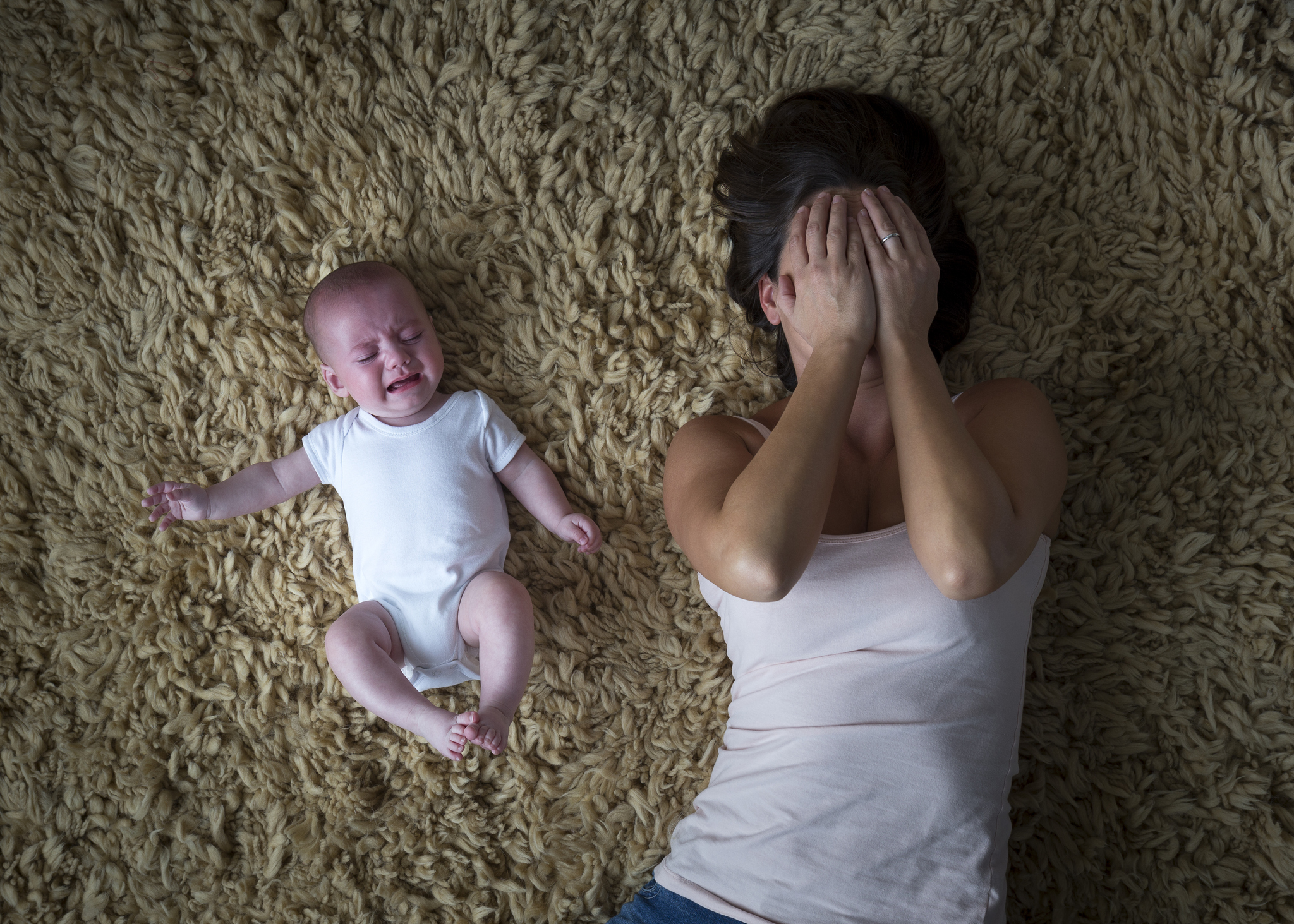 A white-presenting baby in a white onsie cries on a shag carpet next to a dark-haired woman with her hands over her face