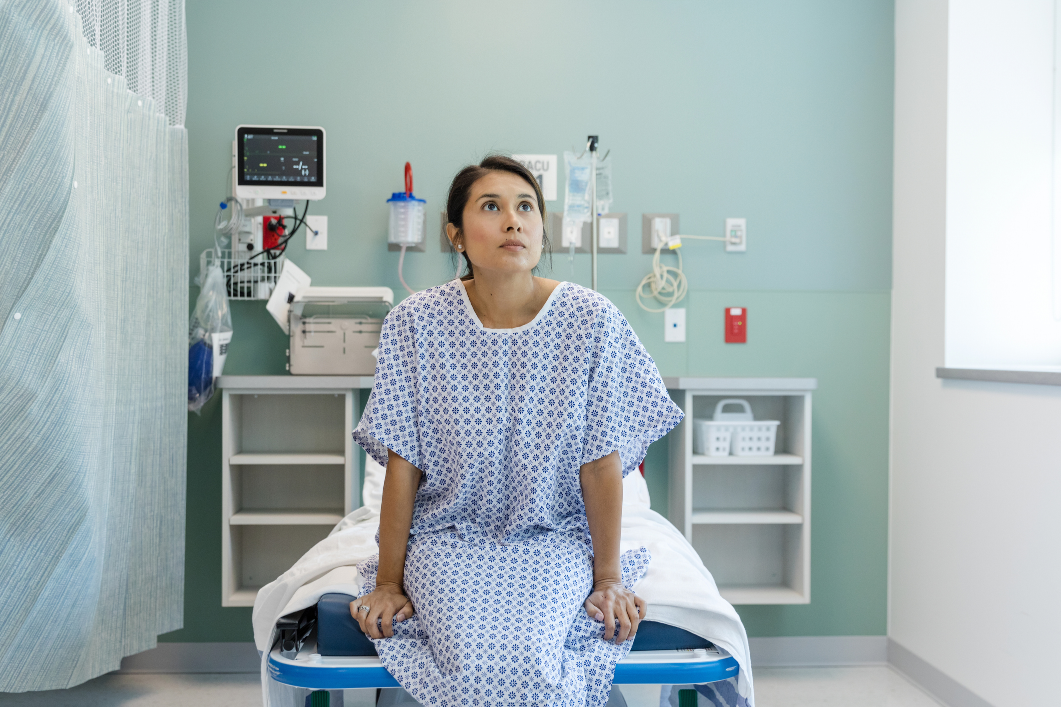 A woman with light brown skin and straight dark hair, wearing a medical gown, sits on the edge of hospital bed and looks nervously upward