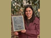 A smiling white-presenting woman with brown hair stands in front of a leafy background holding a plaque