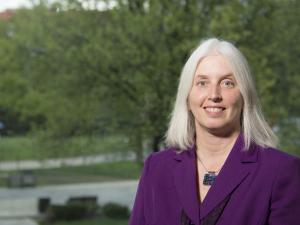 Susan Wood, a white woman with grey hair dressed in a purple suit, stands smiling in front of a tree