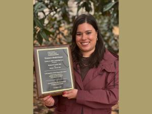 A smiling white-presenting woman with brown hair stands in front of a leafy background holding a plaque