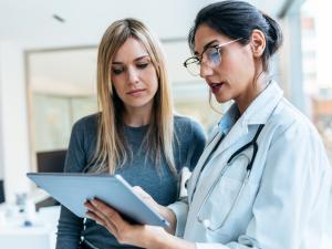 A dark-haired white-presenting woman in a white coat shows something on a clipboard to a blond-haired woman standing next to her
