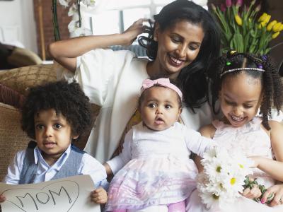 Woman on couch with three young children, one of whom holds a sign with "Mom" inside a heart
