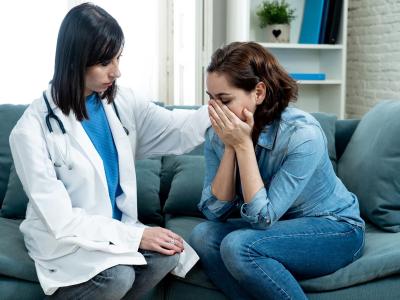 Woman in white coat with stethoscope rests her hand on the shoulder of woman with her head in her hands