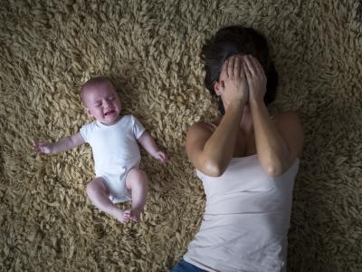 A white-presenting baby in a white onsie cries on a shag carpet next to a dark-haired woman with her hands over her face