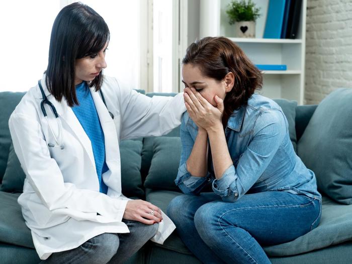 Woman in white coat with stethoscope rests her hand on the shoulder of woman with her head in her hands