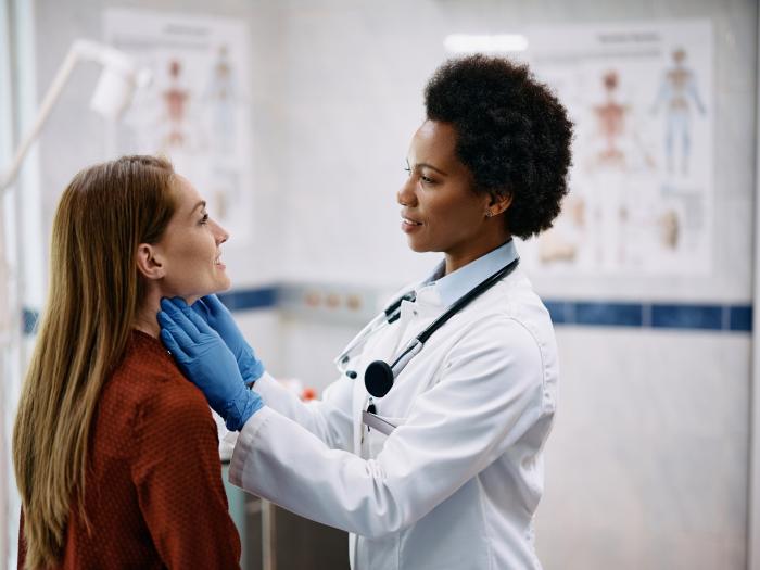 A Black woman in a white coat and blue gloves touches the neck of a white woman with long hair