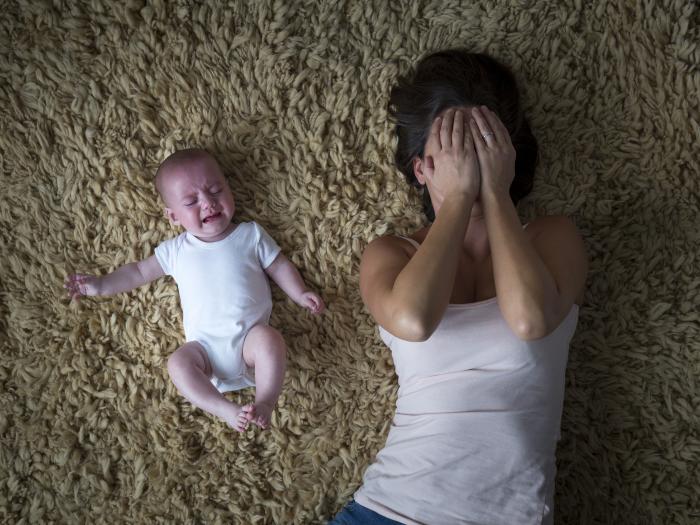 A white-presenting baby in a white onsie cries on a shag carpet next to a dark-haired woman with her hands over her face