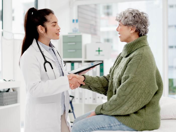 Olive-skinned woman in a white coat with a dark hair in a ponytail shakes the hand of an older white-presenting woman with curly gray hair who wears a thick green sweater and sits on an exam table.